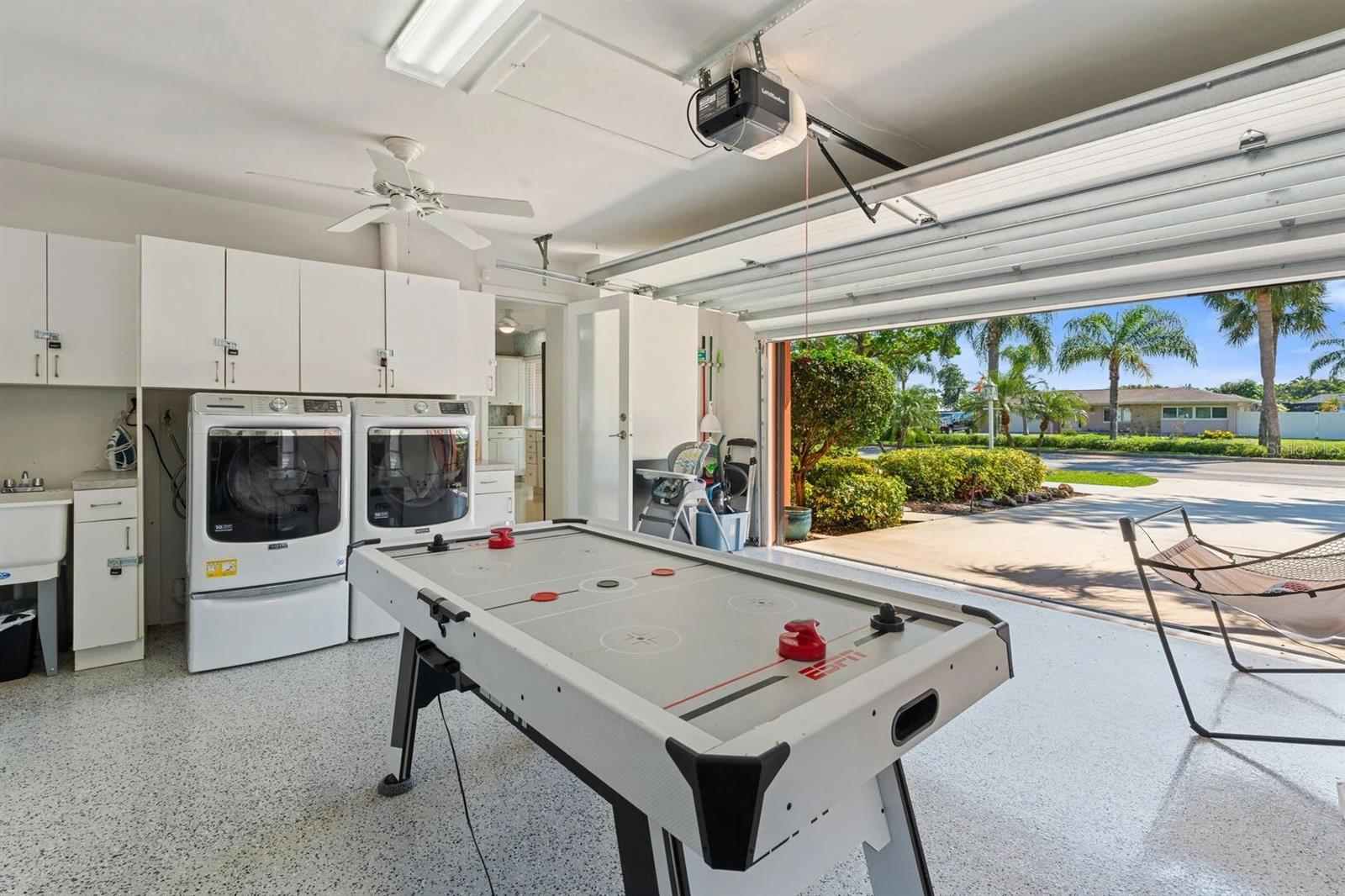 Garage with laundry machines and an air hockey table, open to a palm-lined street.