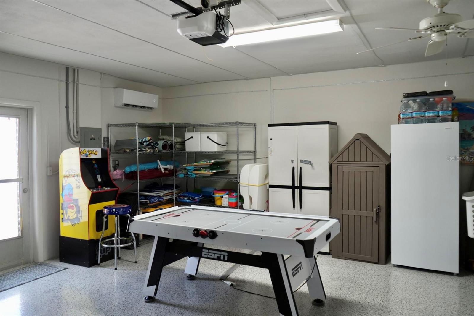 Garage recreation area with an air hockey table, arcade game, storage shelves, and refrigerator.