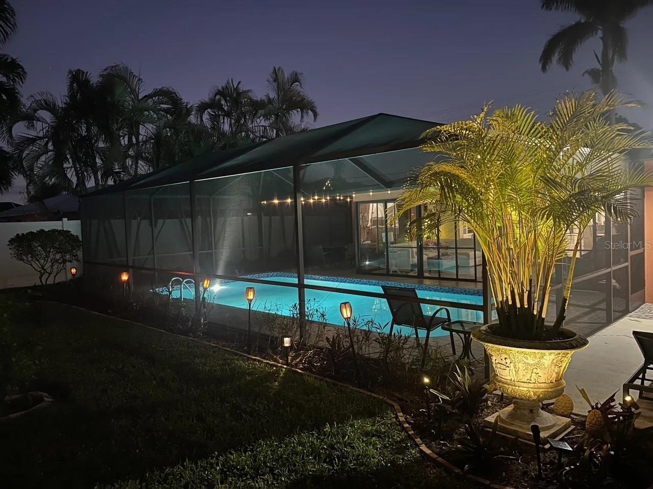Screened pool and patio at night, illuminated by pool and garden lights beneath tropical palms.