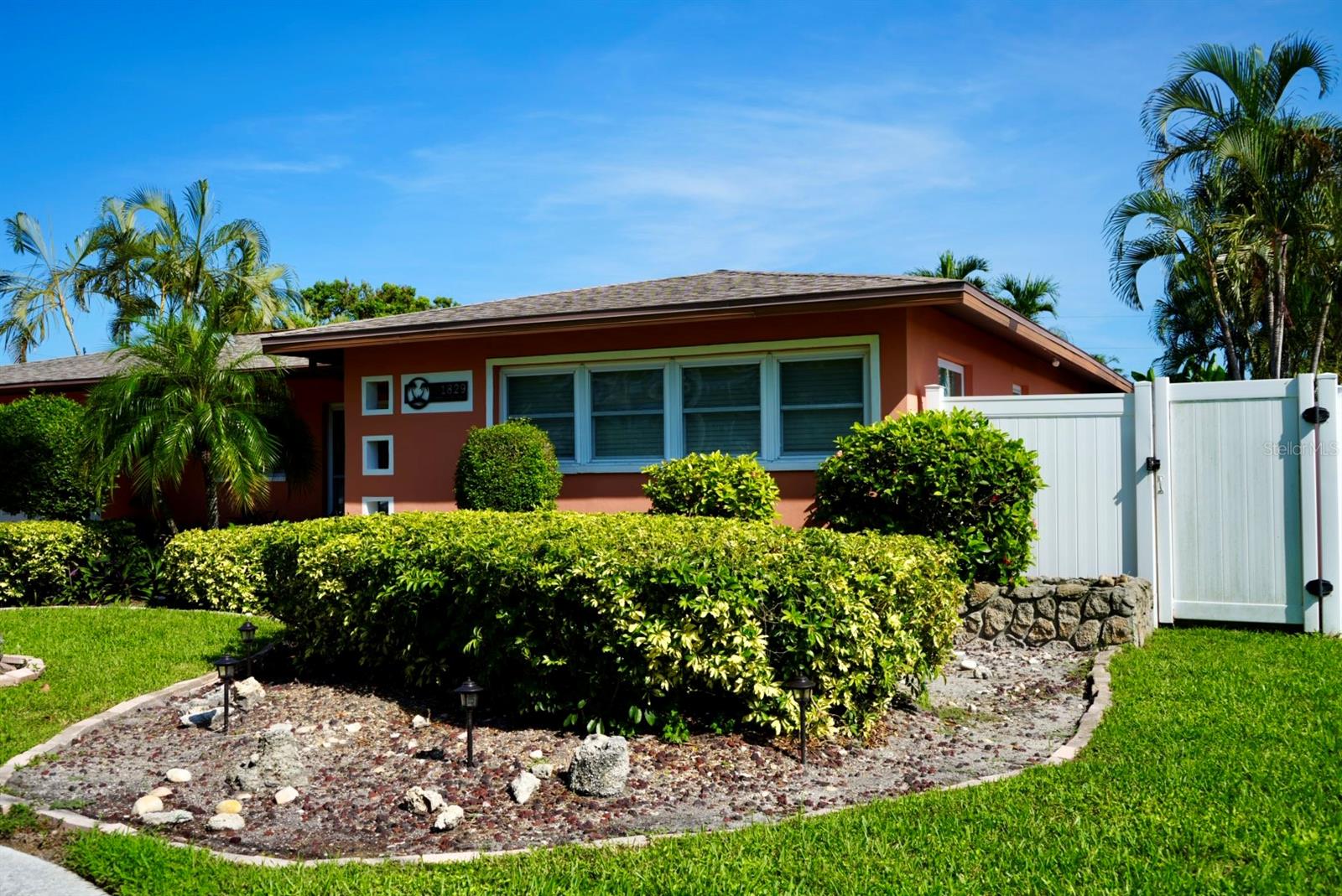 Coral-colored home with a white gate, trimmed hedges, and tropical palms.