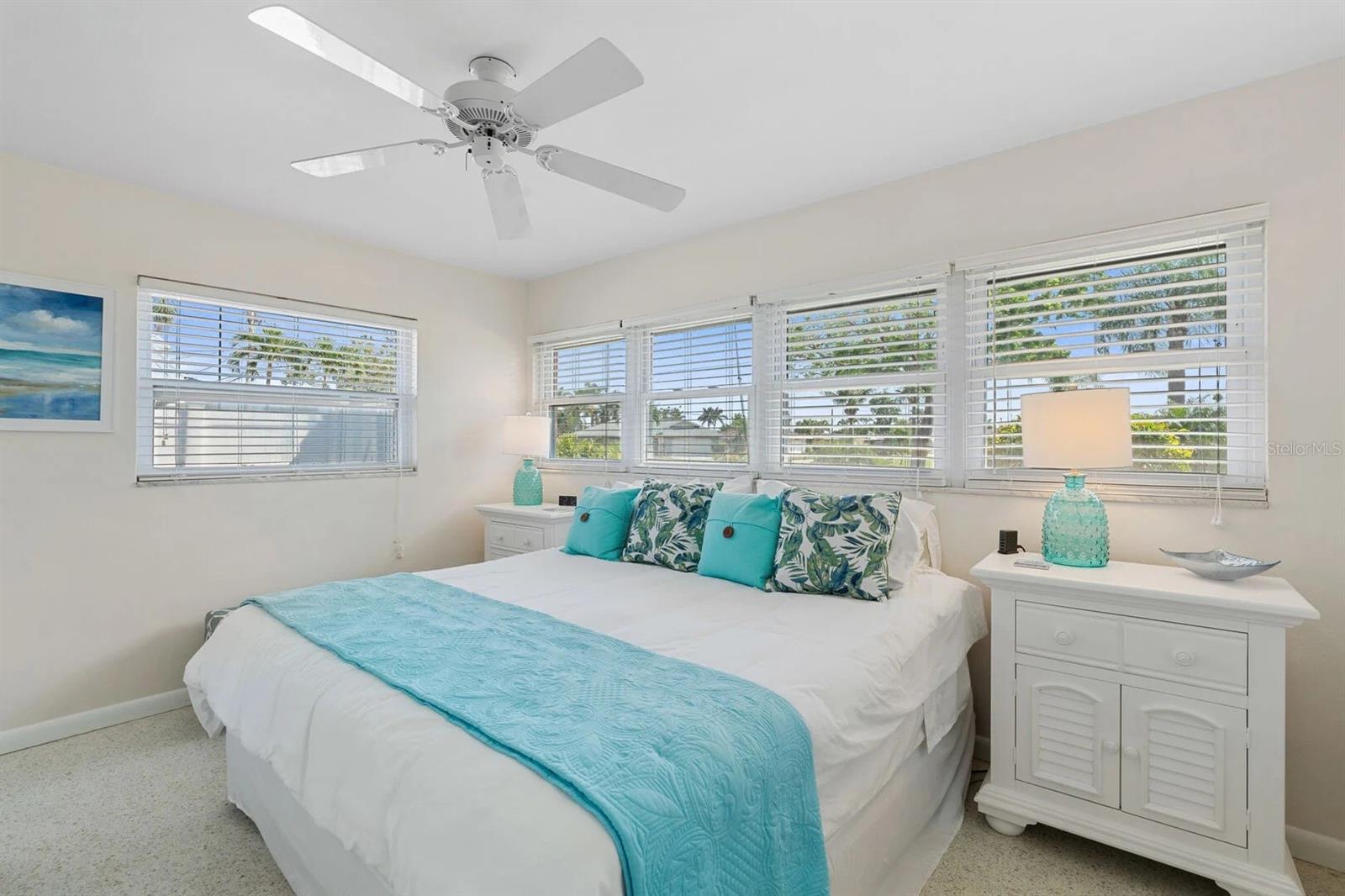Bright bedroom with a white bed, turquoise bedding, ceiling fan, and wide windows with blinds.