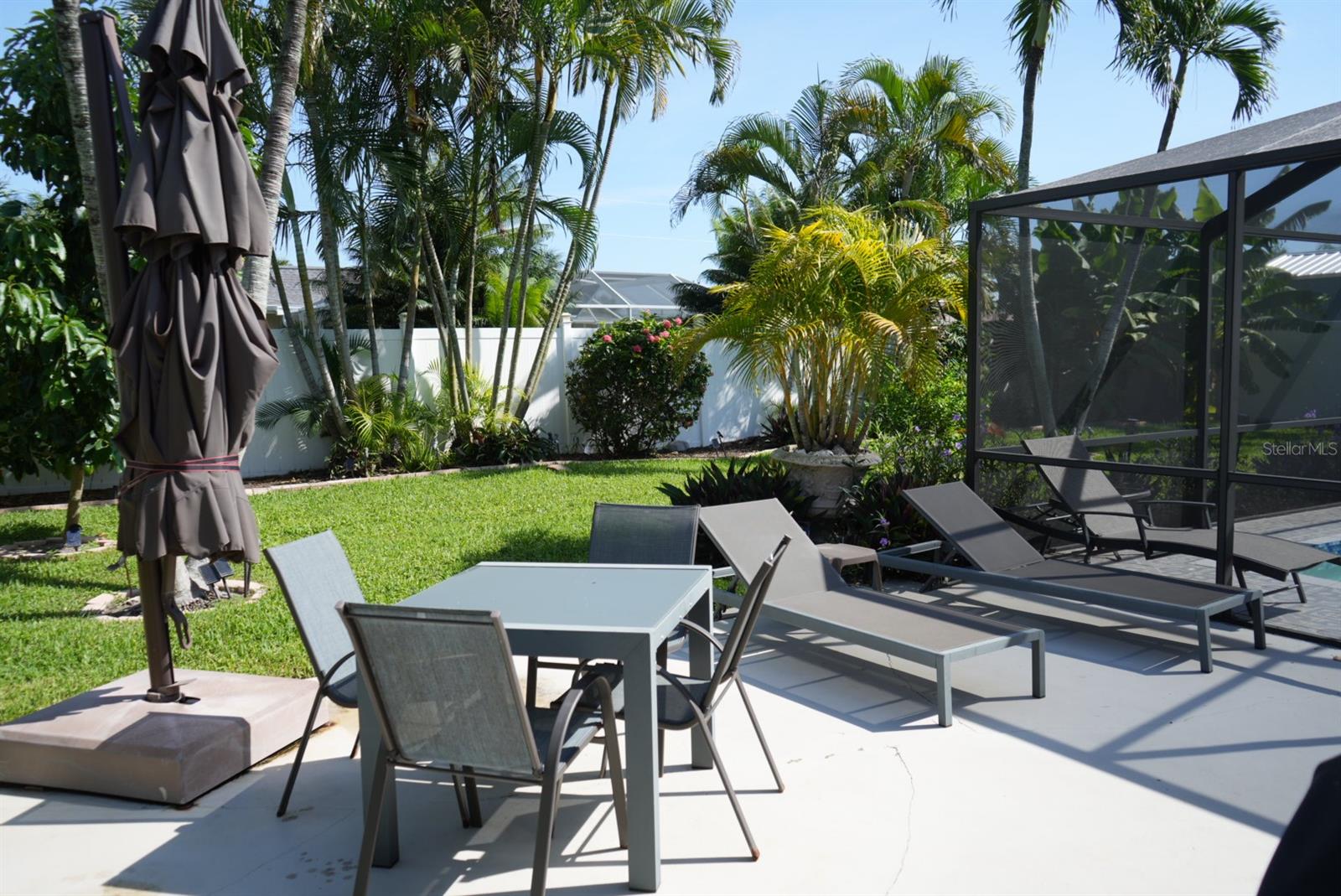 Backyard patio with lounge chairs, outdoor dining set, screened enclosure, and tropical landscaping.