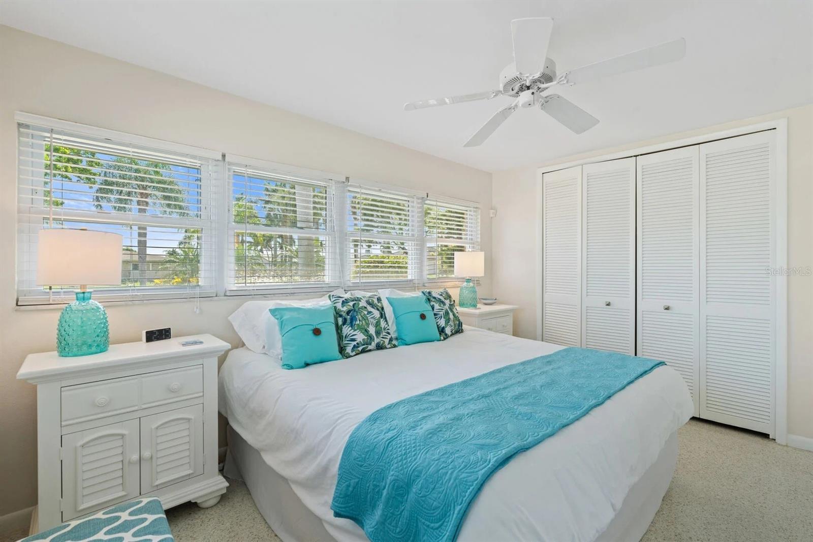 Bedroom with turquoise bedding, louvered closet doors, and wide windows overlooking palm trees.
