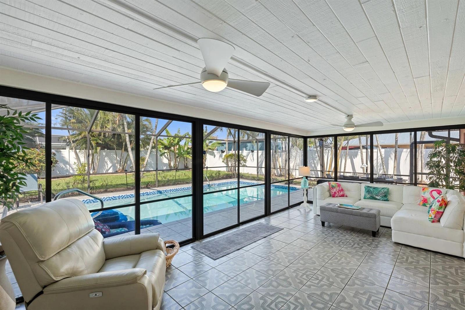 Enclosed sunroom with white seating and ceiling fans overlooking a backyard pool.
