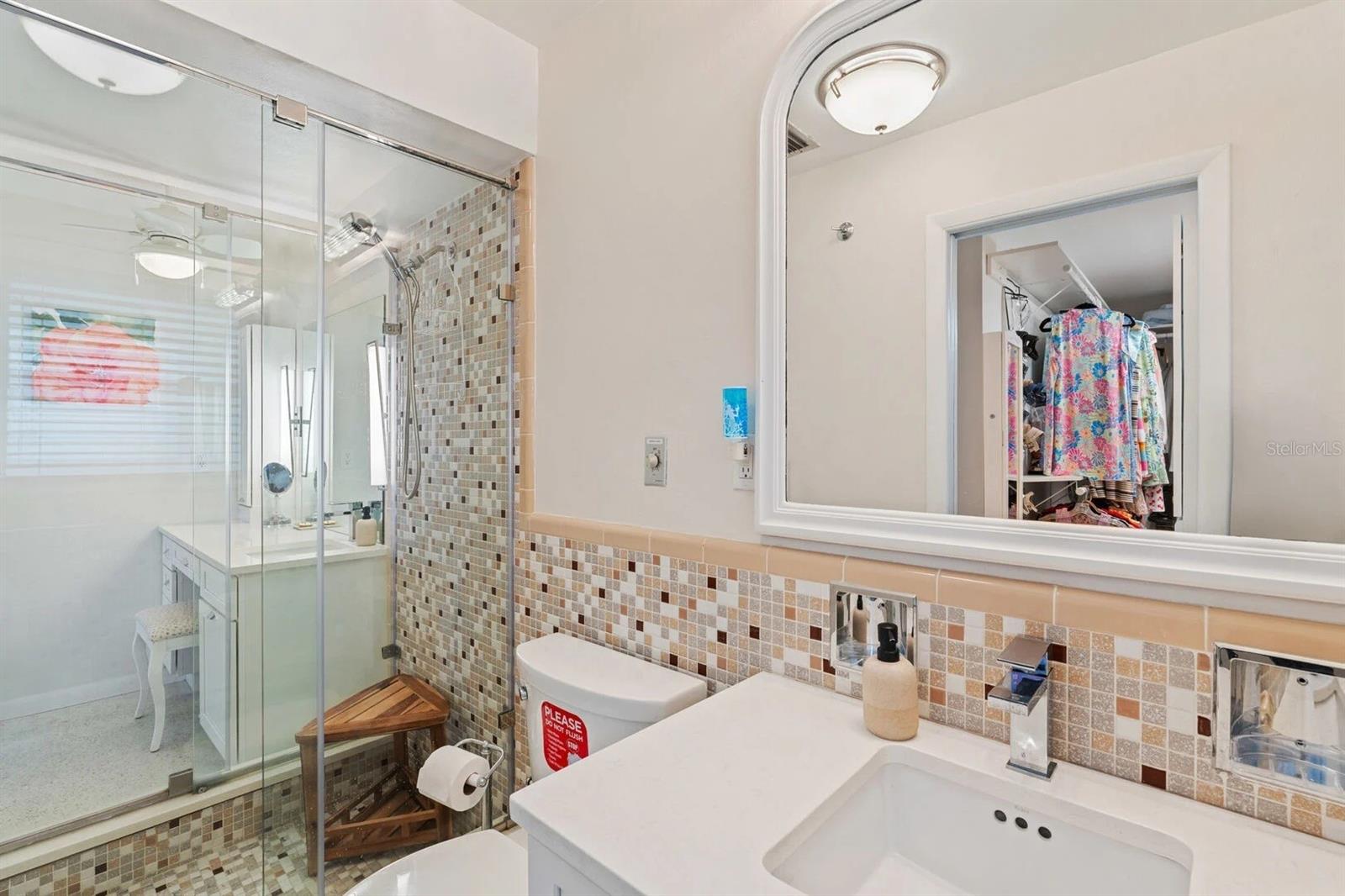 Bathroom with a glass shower, mosaic tile, white vanity, and mirror reflecting an open closet.