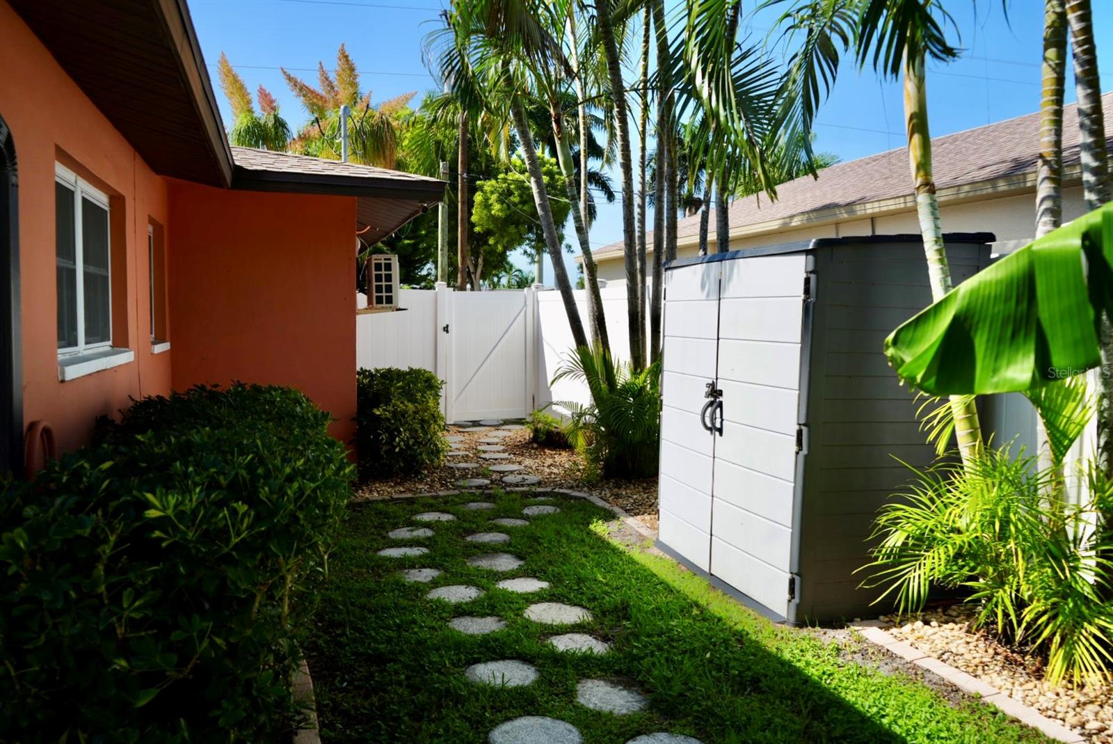 Side yard with a gray storage shed, white fence, tropical palms, and stepping-stone path.