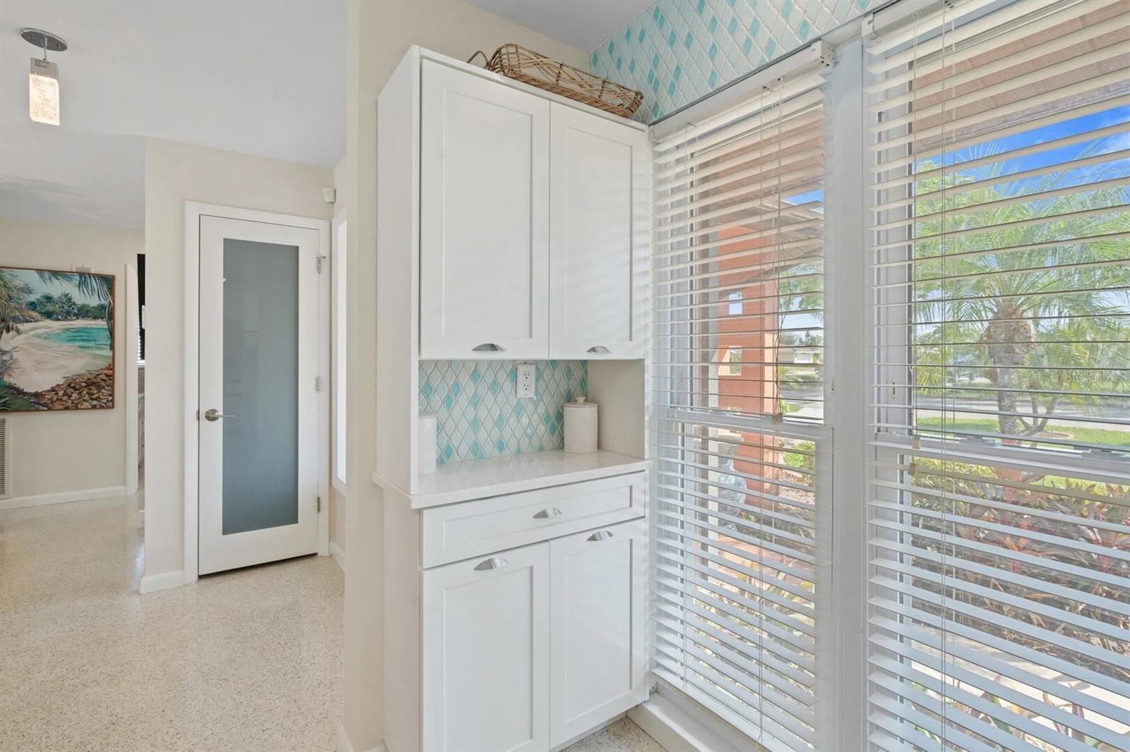 White built-in kitchen cabinets and counter beside tall windows overlooking palm trees.