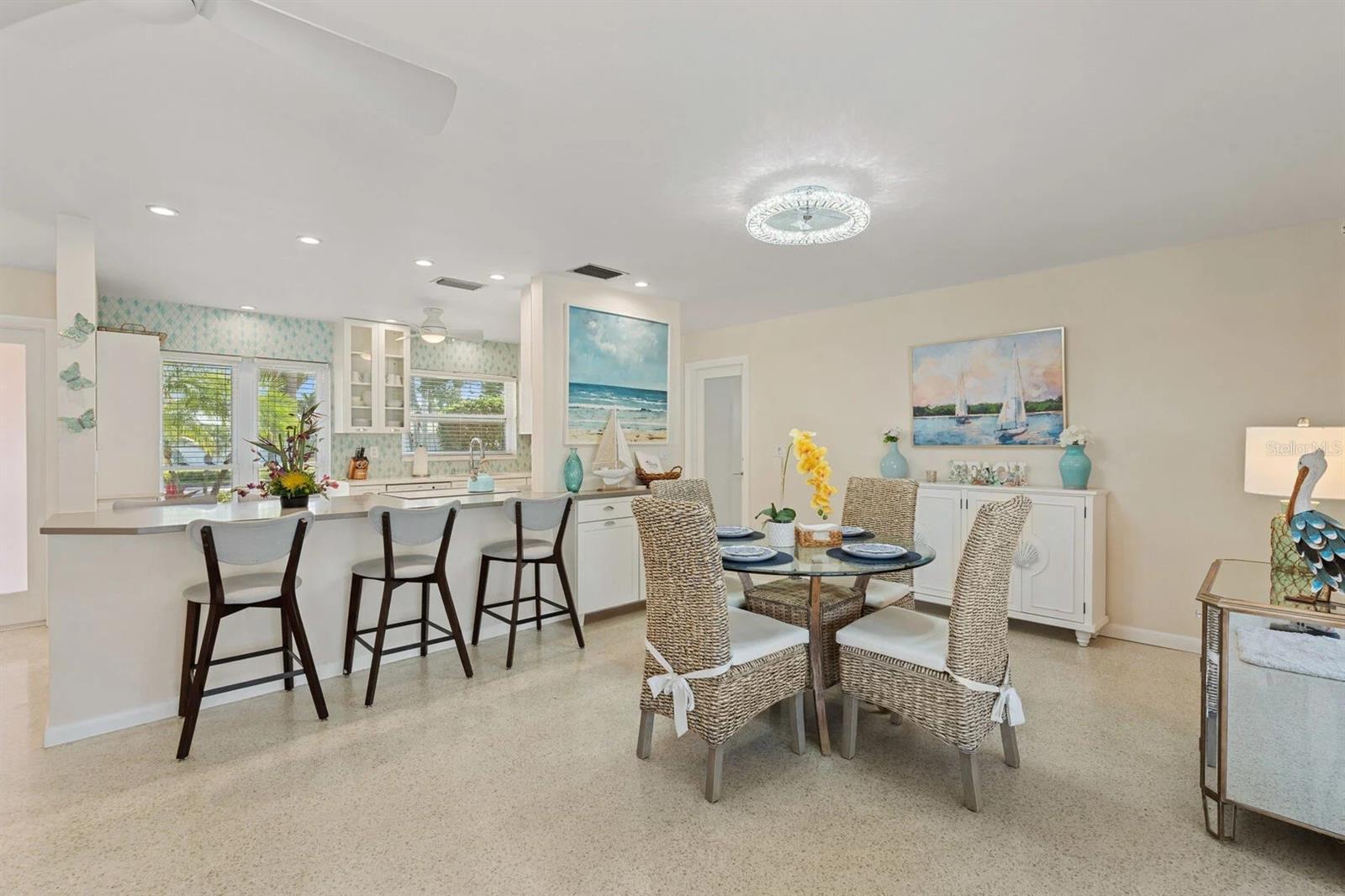 Open kitchen and dining area with a white breakfast bar, woven chairs, and coastal artwork.
