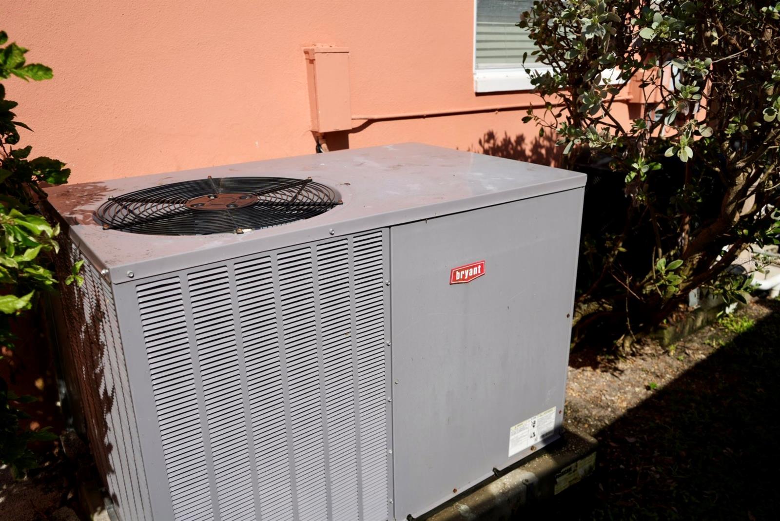 Outdoor air-conditioning condenser beside a coral stucco wall and shrubs.