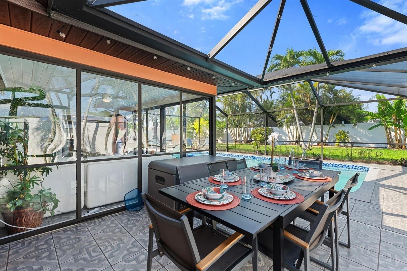 Outdoor dining table on a screened lanai beside a pool, with tropical landscaping beyond.