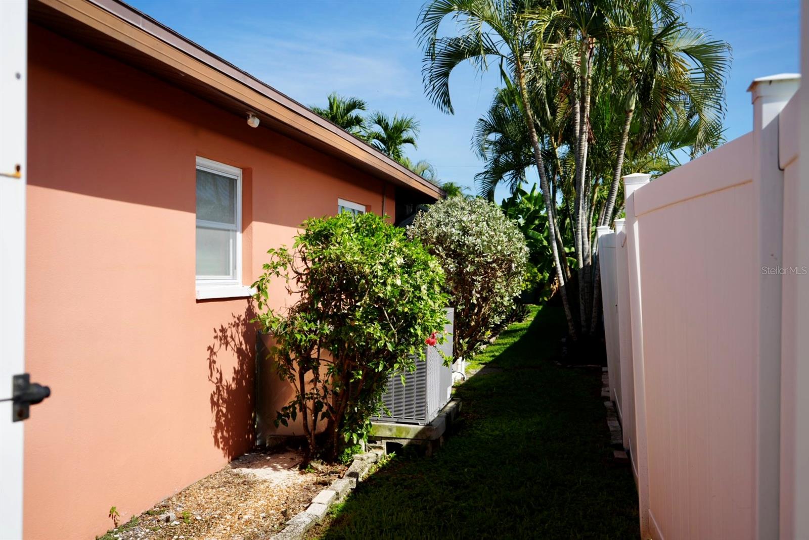 Coral stucco home beside a narrow grassy yard bordered by a white fence and tropical palms.