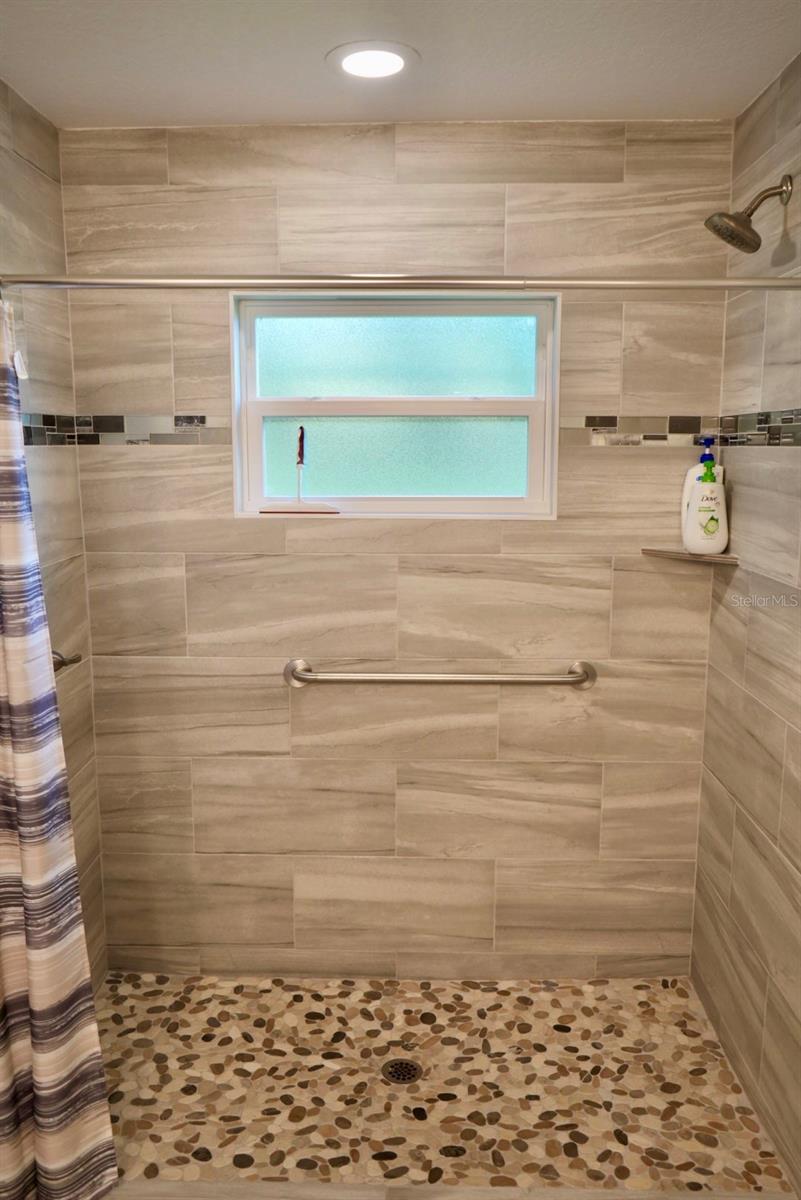 Walk-in shower with beige tile, a pebble-stone floor, frosted window, and metal grab bar.