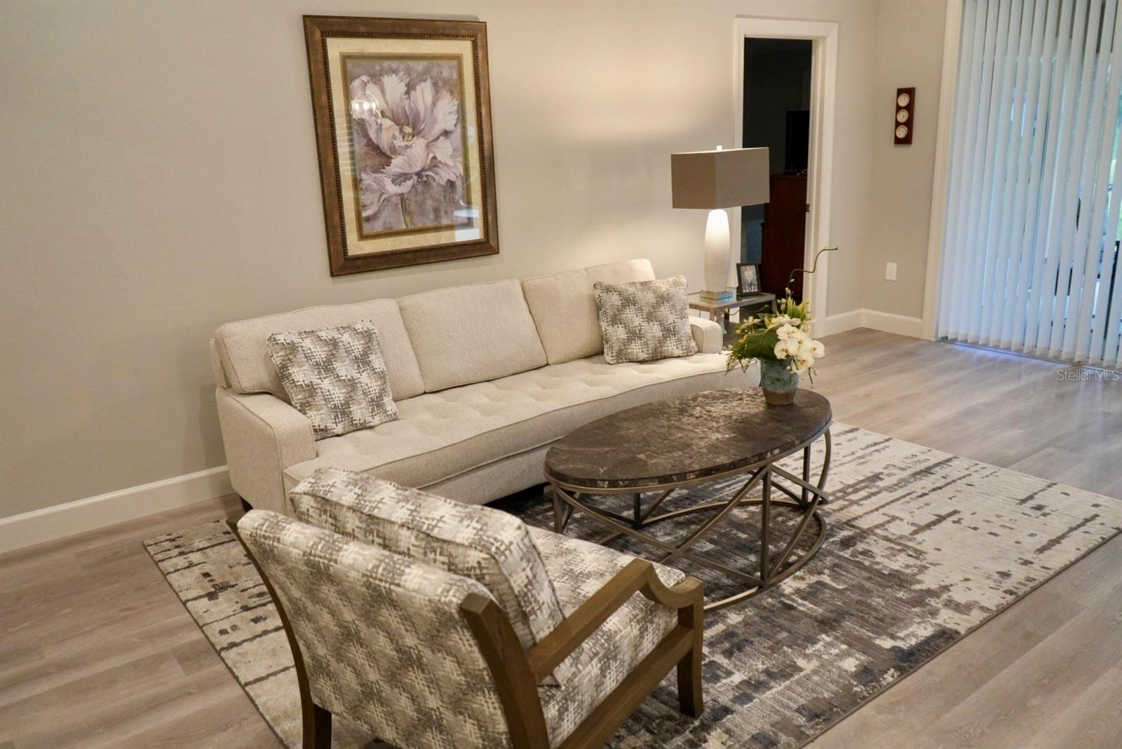 Furnished living room with a cream sofa, patterned armchair, oval coffee table, and sliding blinds.