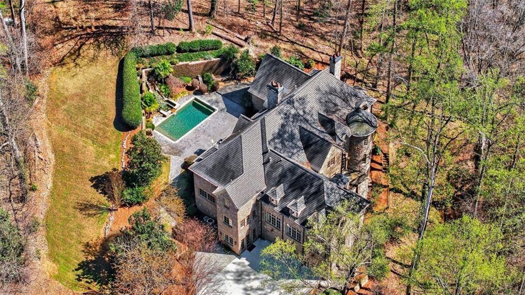 Aerial view of a stone home with a rectangular pool, patio, and wooded grounds.