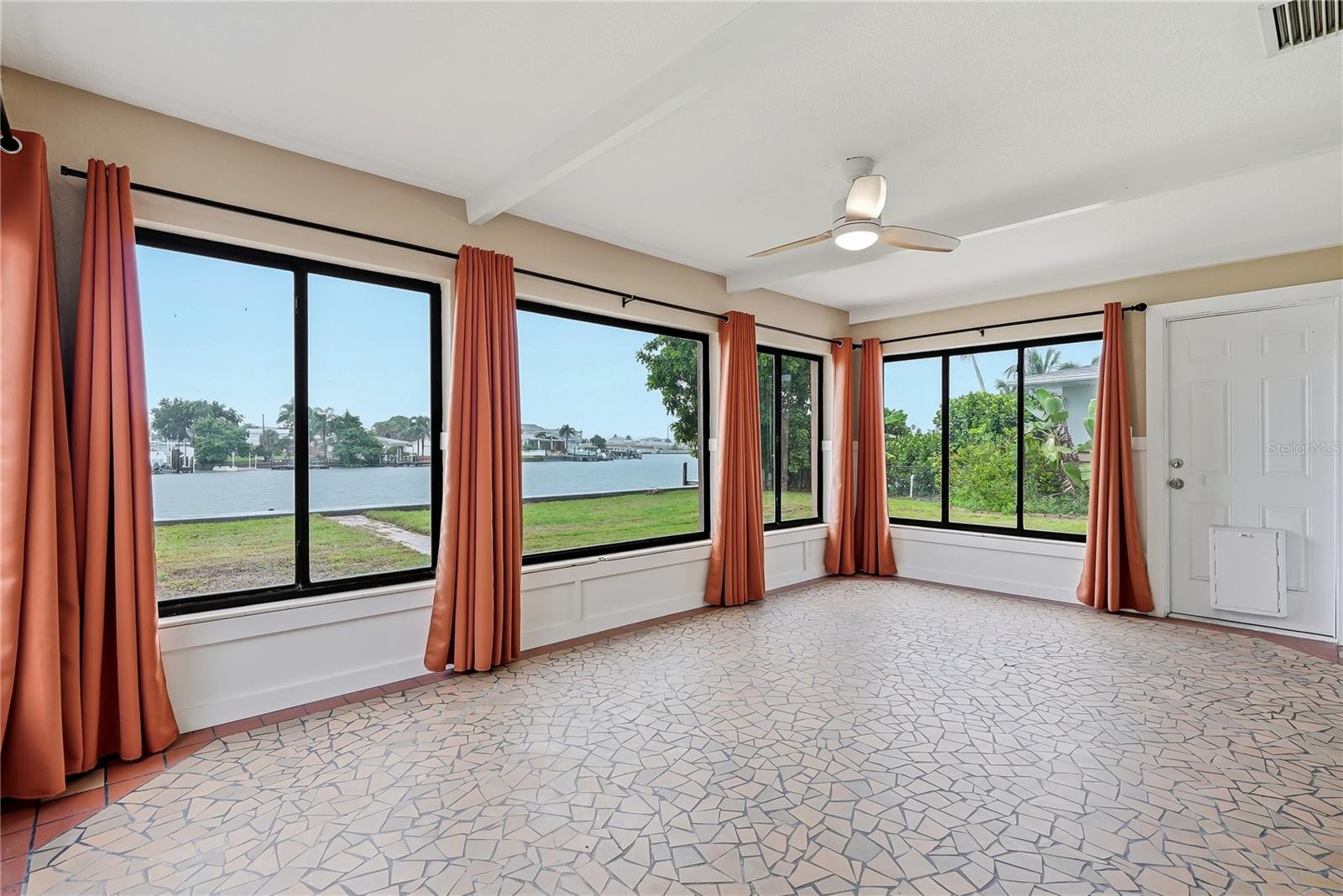 Empty waterfront room with coral curtains, large windows overlooking a canal, and patterned tile flooring.