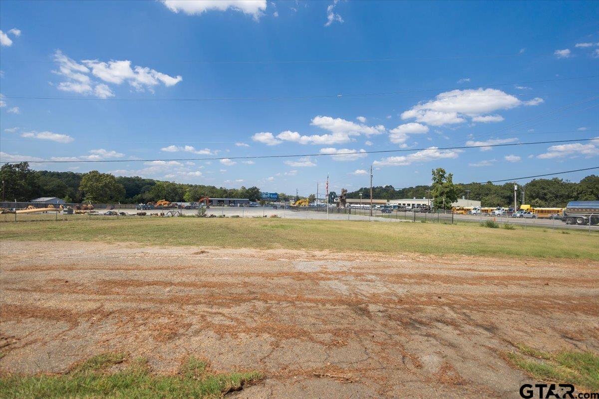 Vacant lot with a gravel foreground and grassy field, with commercial buildings beyond a fence.