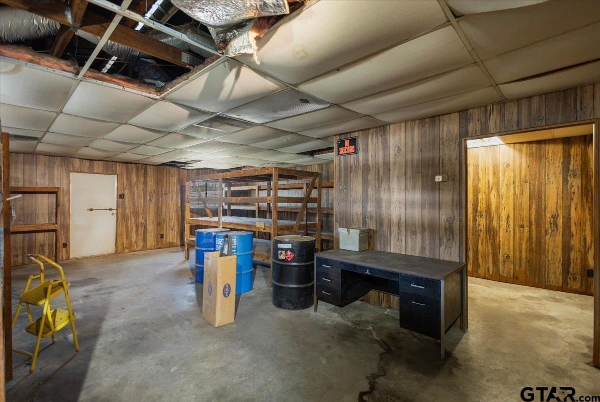 Storage room with wood-paneled walls, shelving, drums, a desk, and a damaged ceiling.