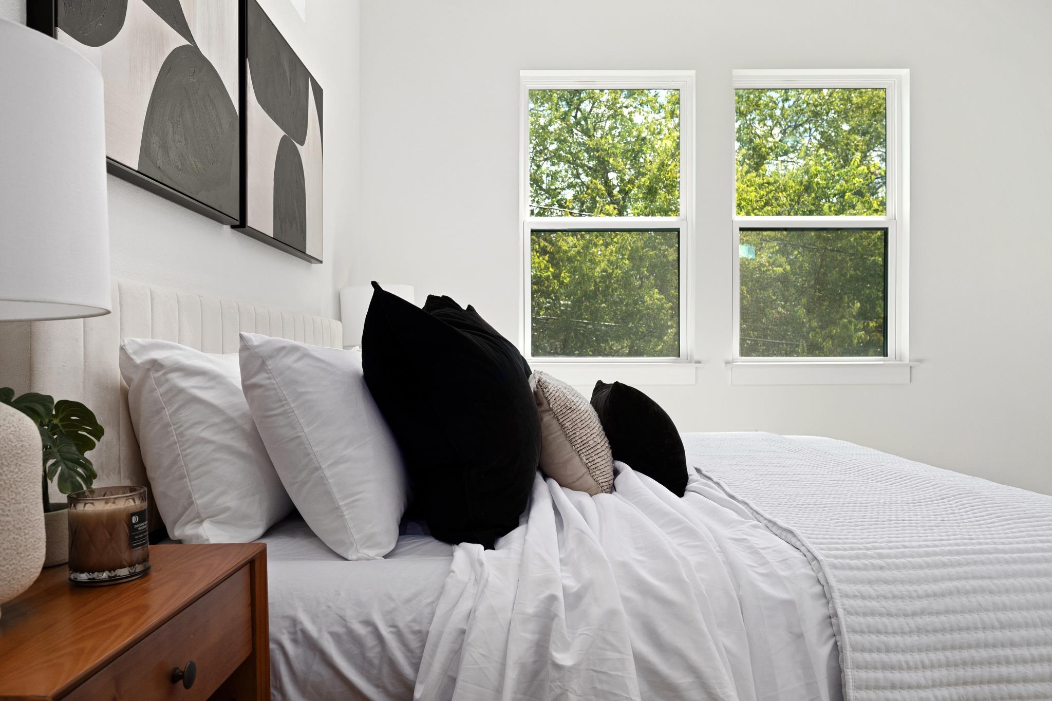 Bright bedroom with a white bed, black accent pillows, and two windows overlooking leafy trees.