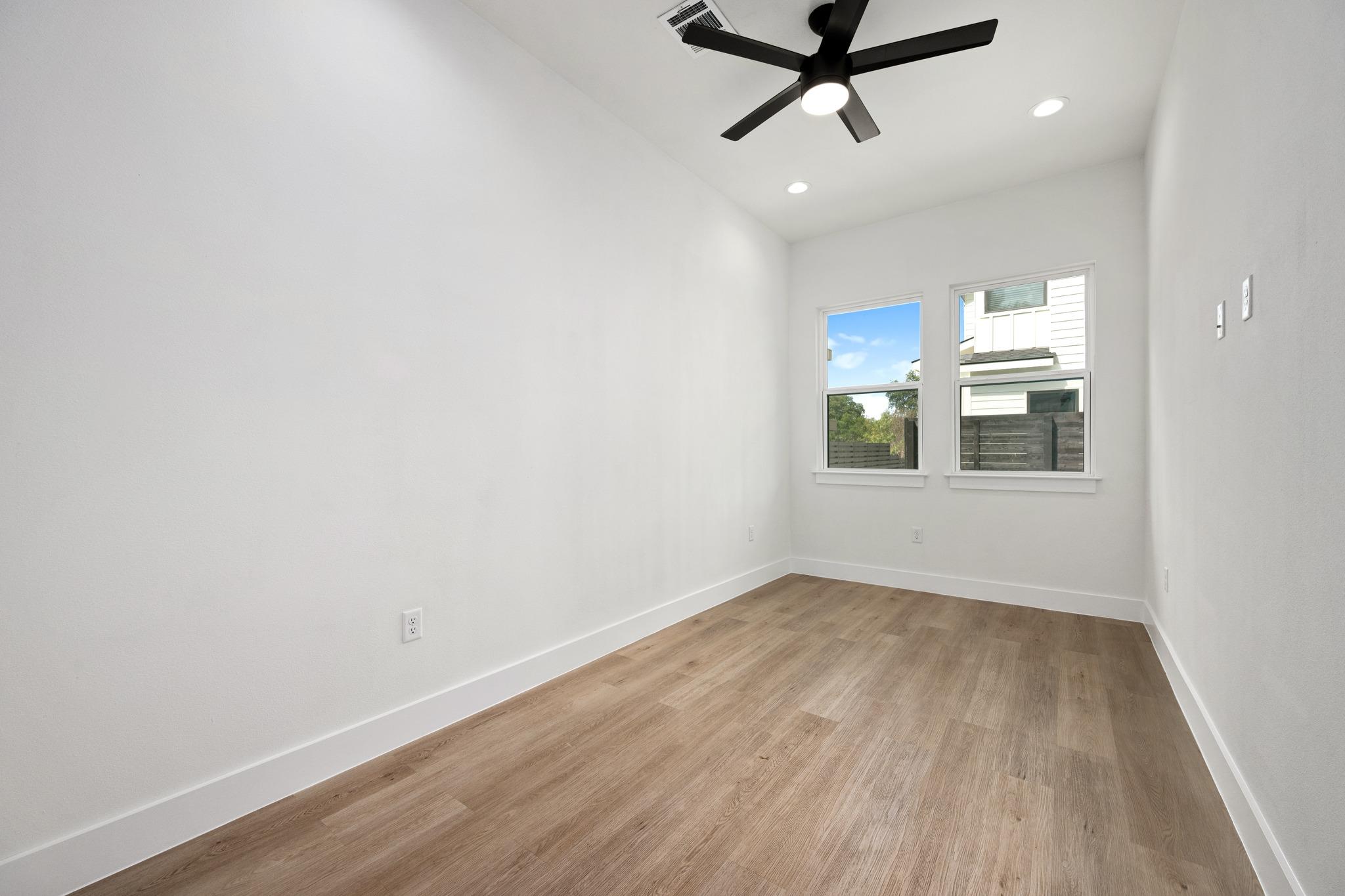Empty room with wood-look floors, a black ceiling fan, and two windows overlooking a fence.