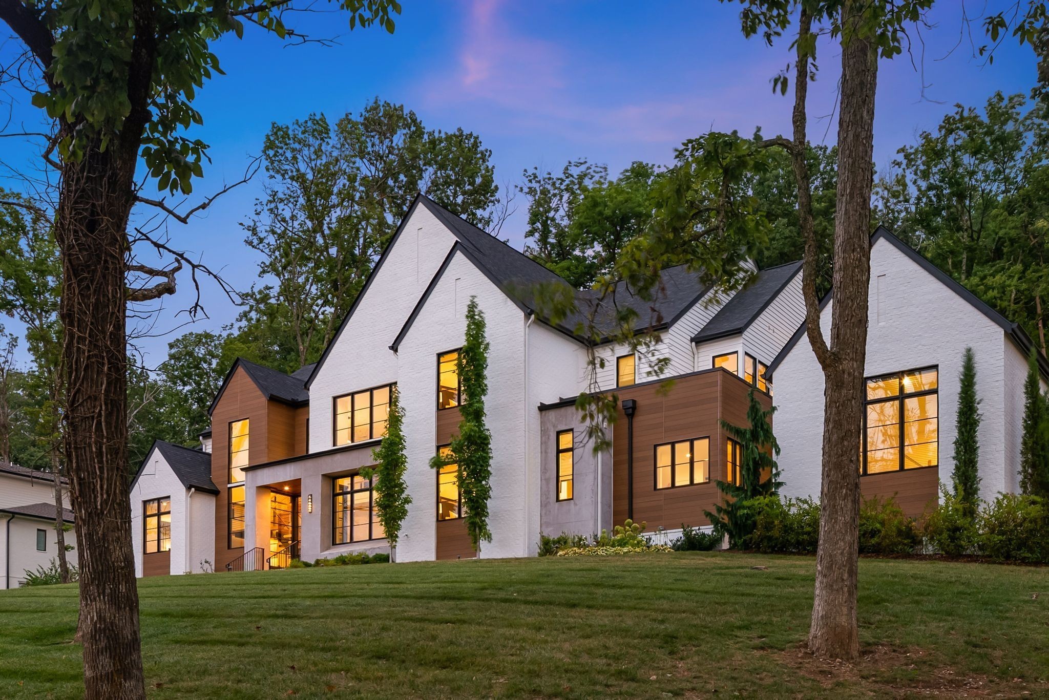 Modern white-and-brown house with steep dark roofs and warmly lit windows among mature trees at dusk.