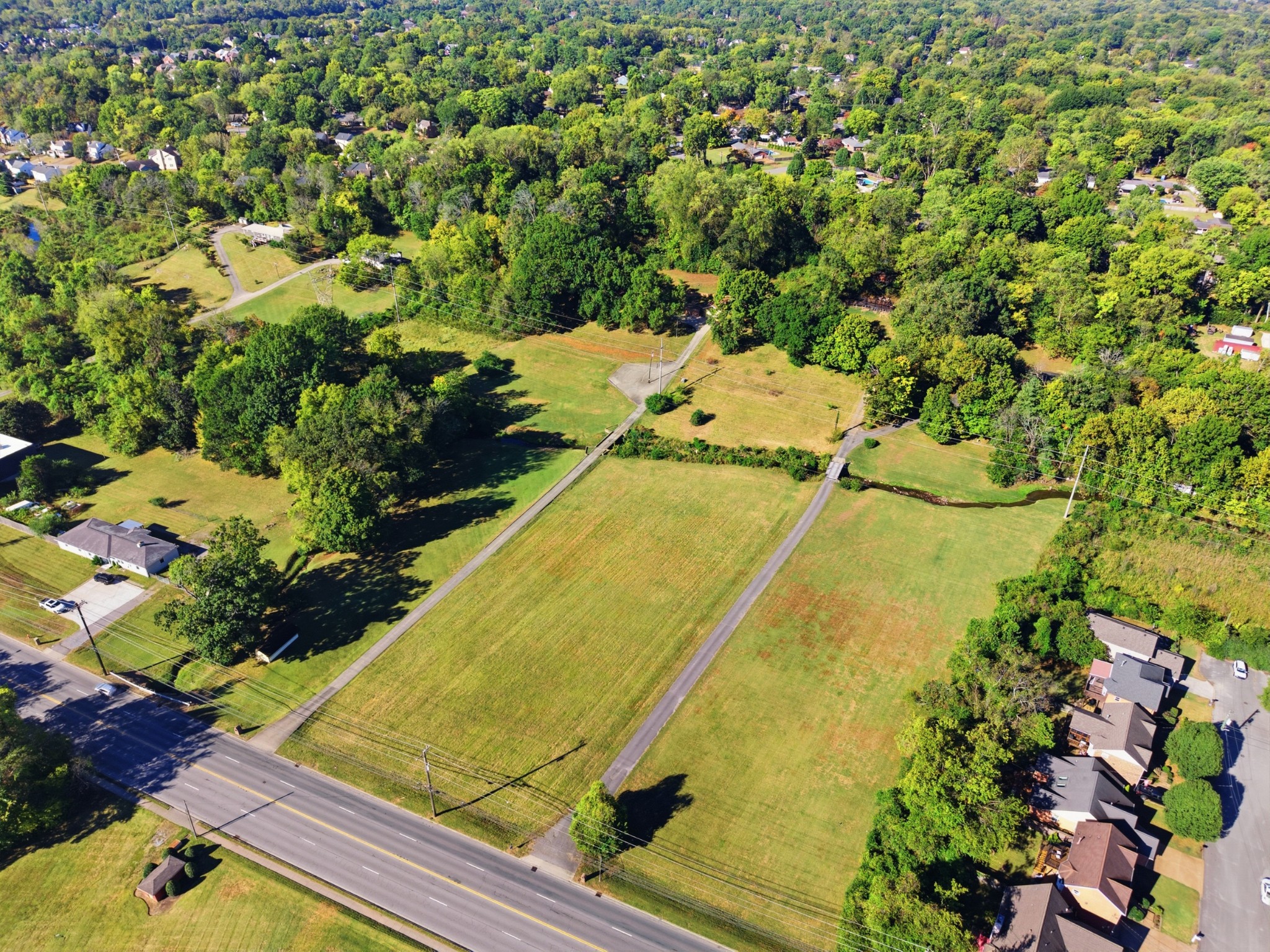 Aerial view of a grassy vacant parcel with paved access lanes, wooded edges, and nearby homes.