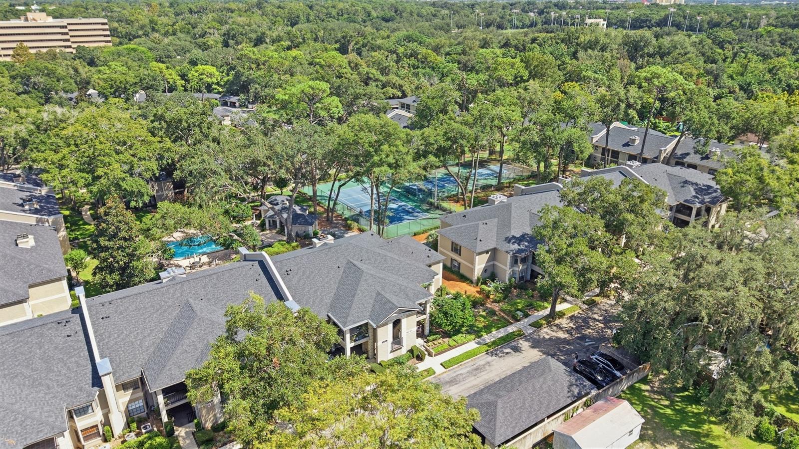 Aerial view of residential buildings, swimming pools, and tennis courts among mature trees.