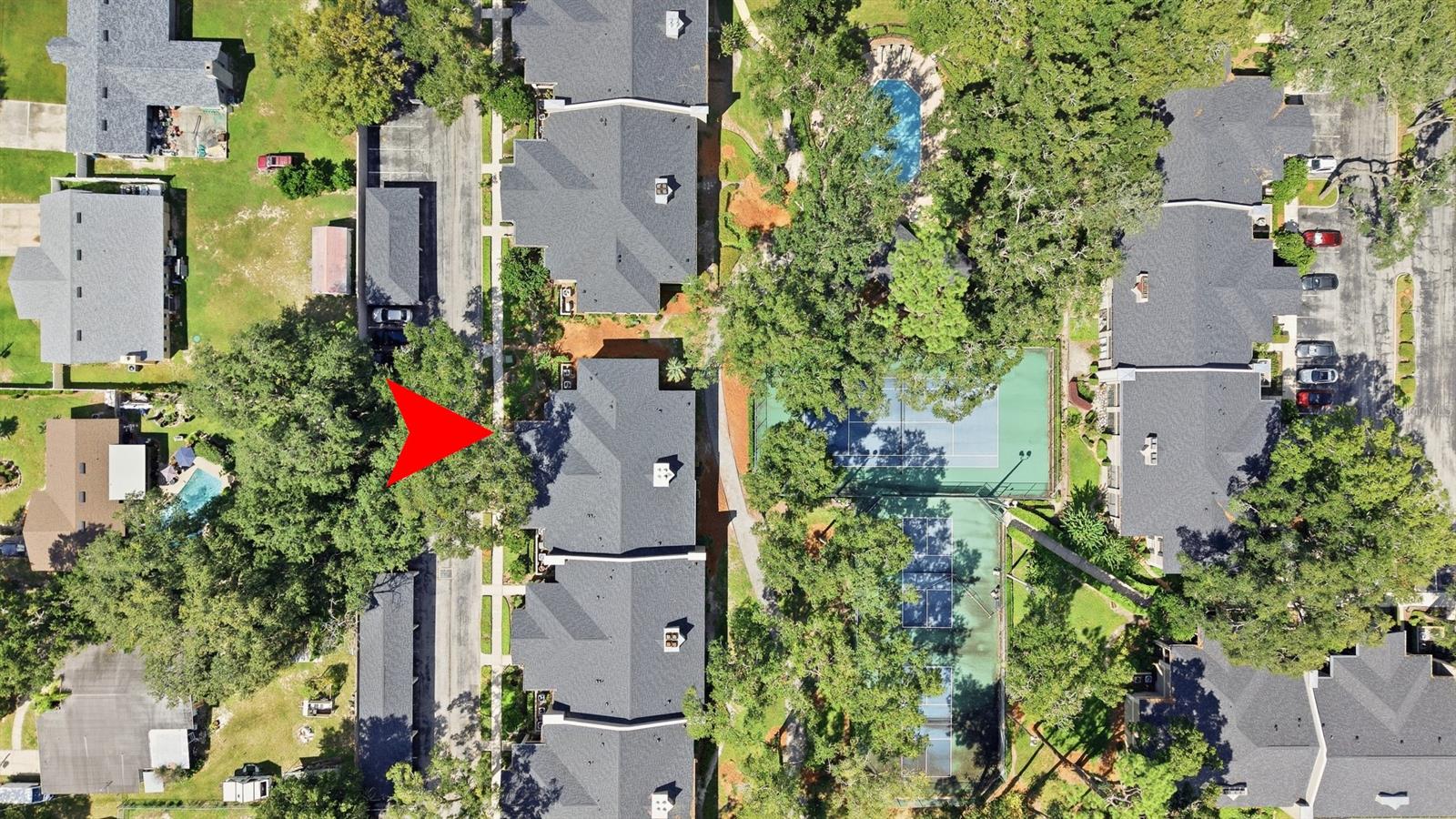 Aerial view of a condominium building indicated by a red arrow, beside tree-lined tennis courts and a swimming pool.