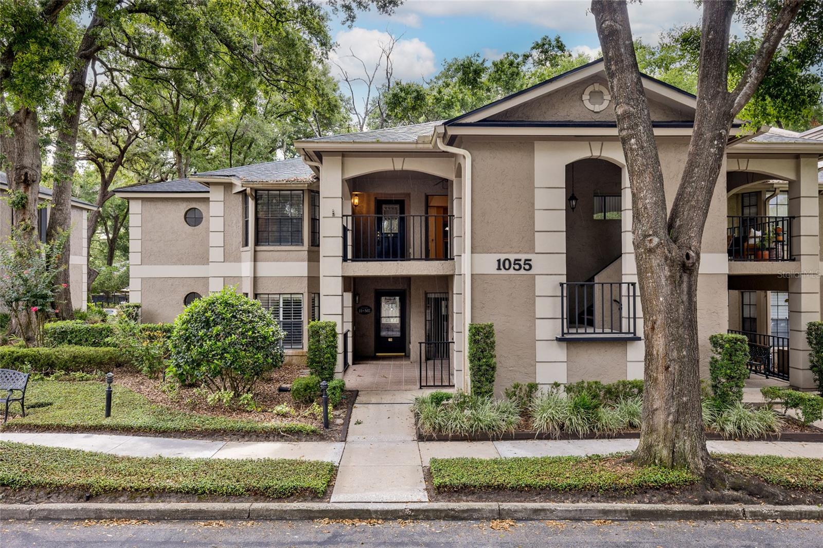 Two-story beige building with covered balconies, black railings, and mature trees.