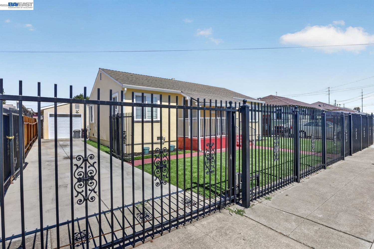 Beige house behind a tall black metal fence, with a driveway, detached garage, and small front lawn.