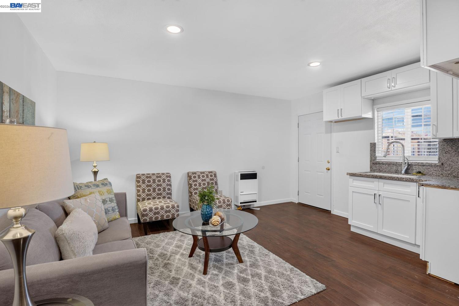 Staged living room with gray sofa and patterned chairs beside a bright kitchen with white cabinets.