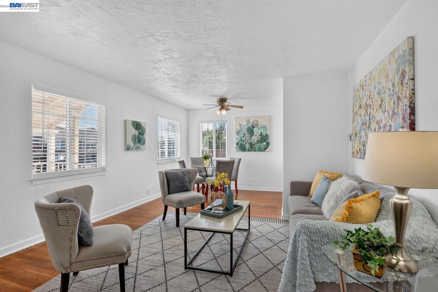 Staged living room with gray seating, hardwood floors, and a dining area by the windows.