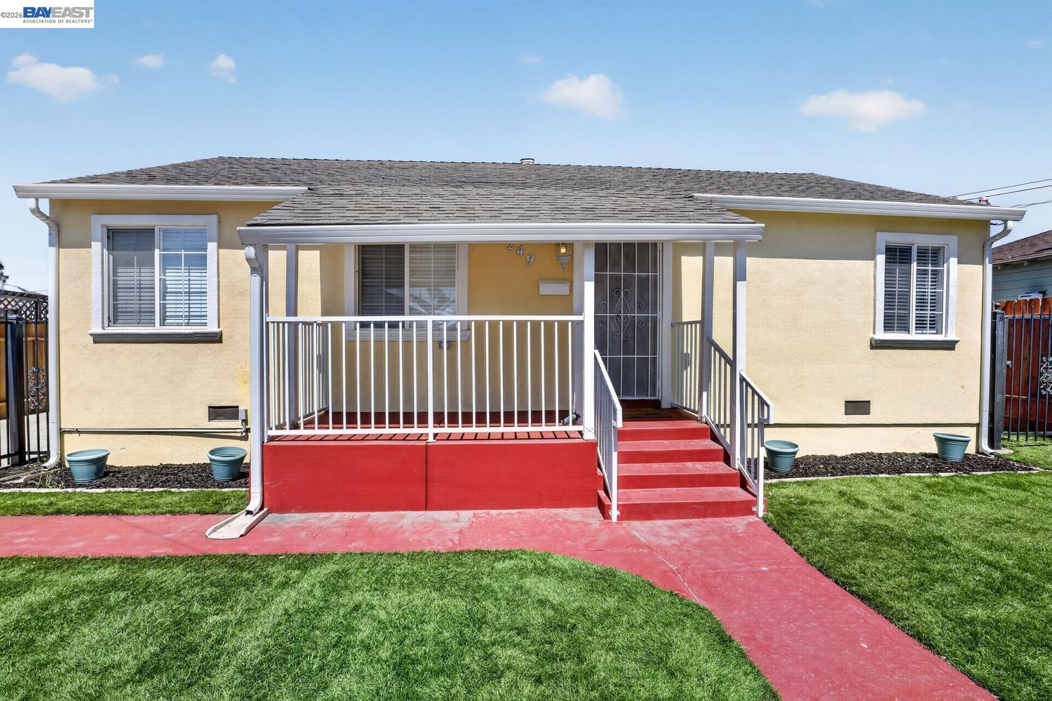 Single-story yellow stucco house with a covered porch, white railings, and a red walkway across the lawn.