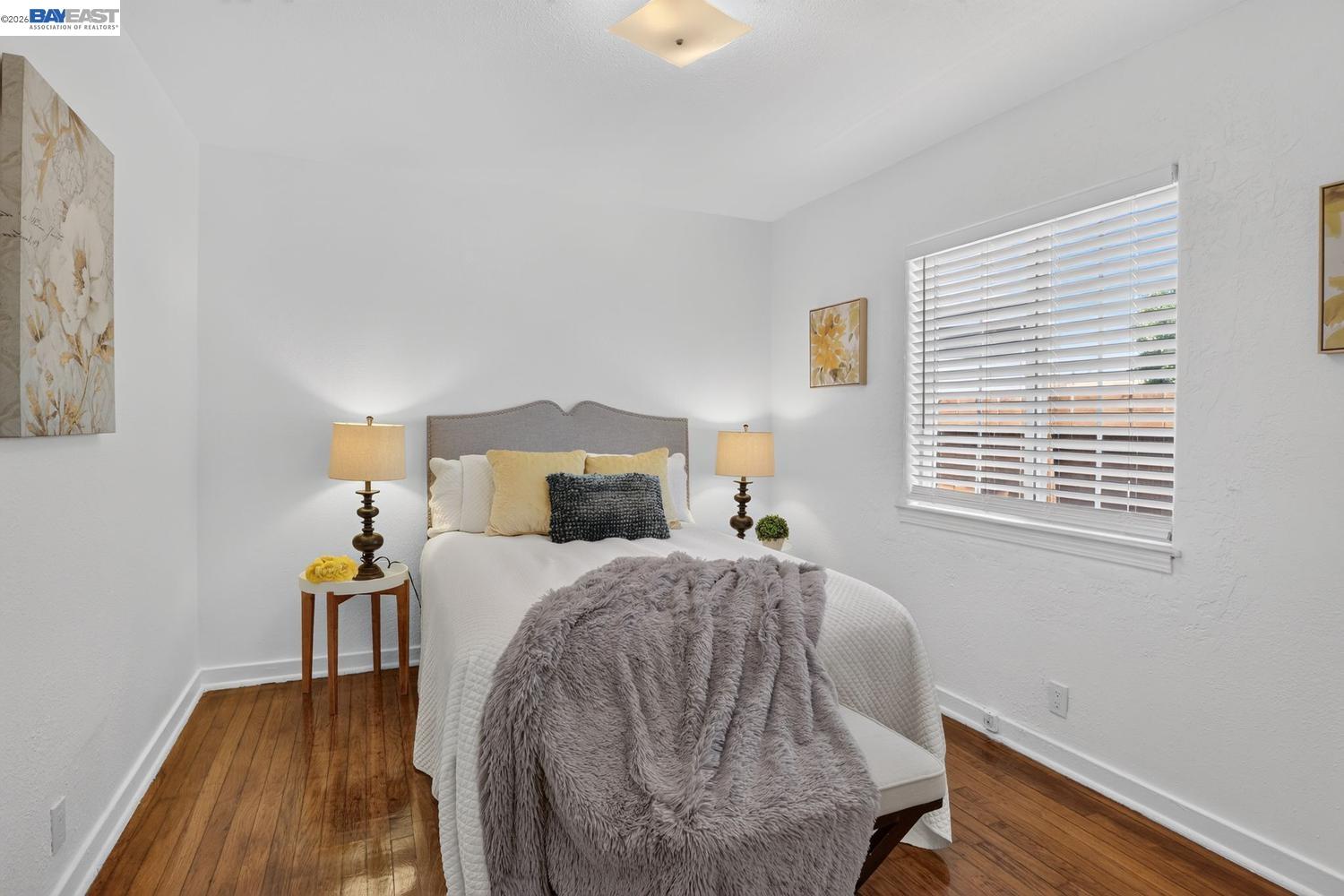 Staged bedroom with a white bed, plush gray throw, and warm wood flooring.