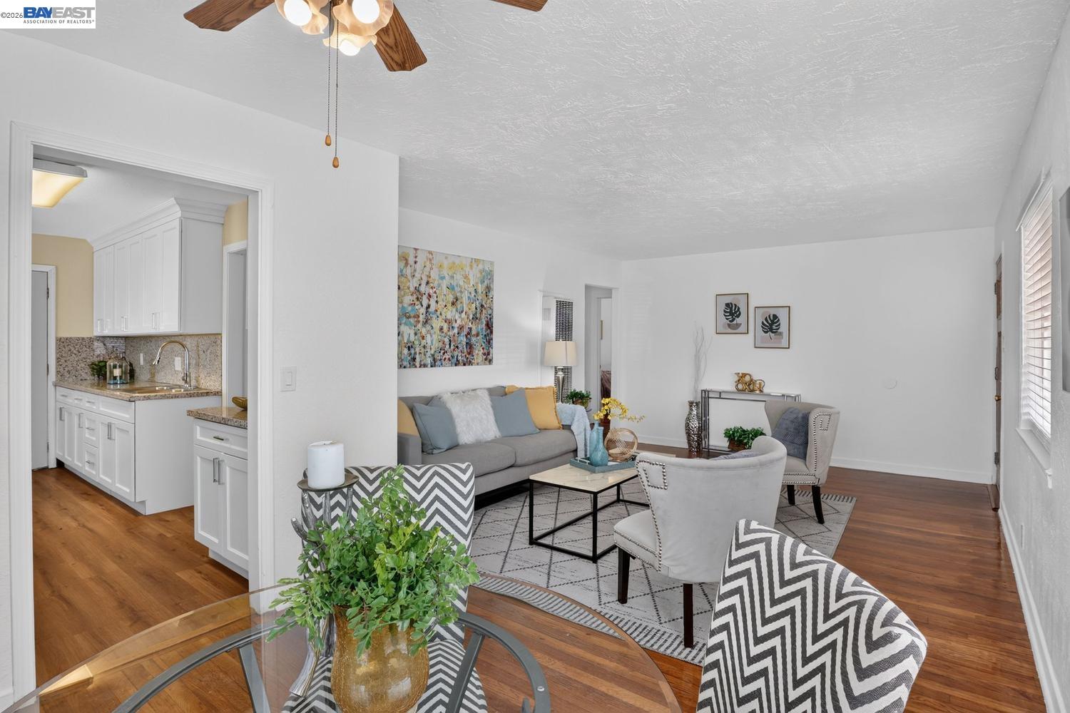 Staged living room with hardwood floors, patterned armchairs, and an adjoining white kitchen.