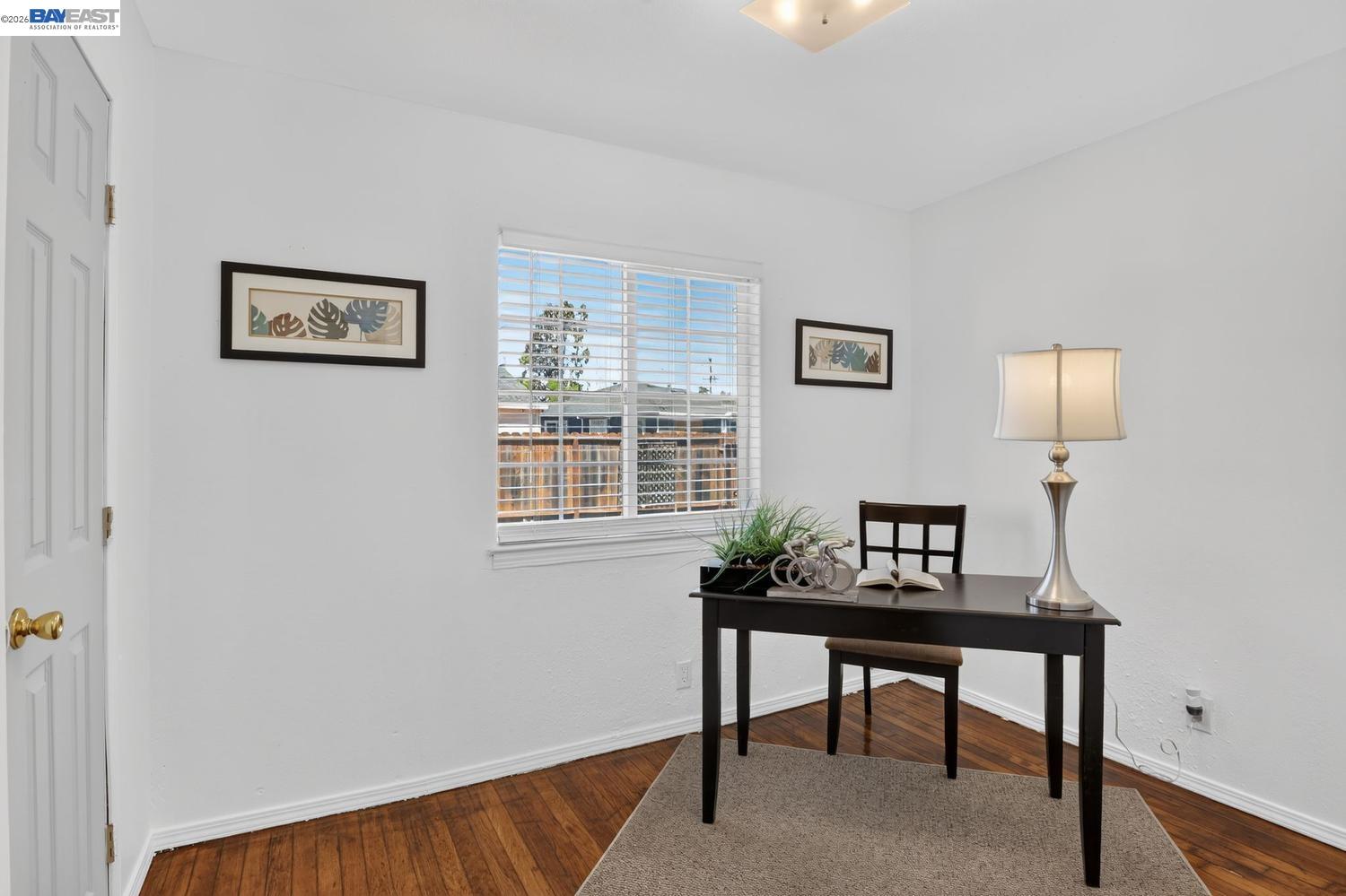 Staged home office with a dark desk, table lamp, botanical prints, and hardwood floors.