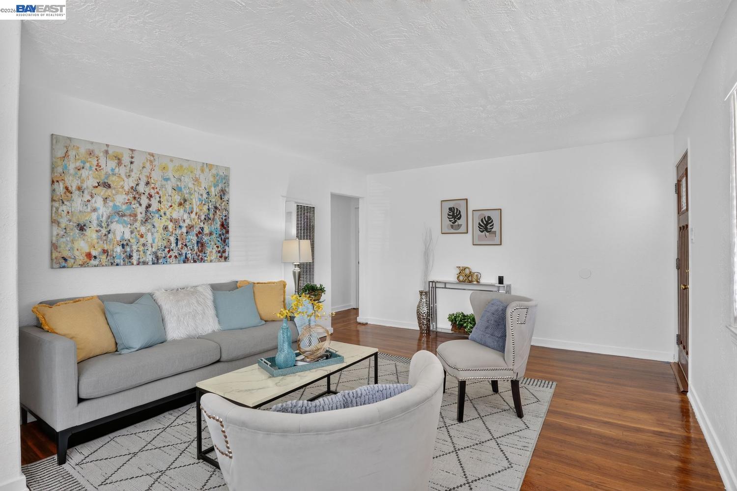 Staged living room with a gray sofa, colorful abstract artwork, white accent chairs, and hardwood floors.