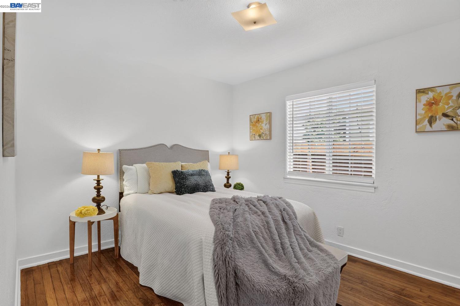 Furnished bedroom with a white bed, gray throw, hardwood floors, and a window with blinds.