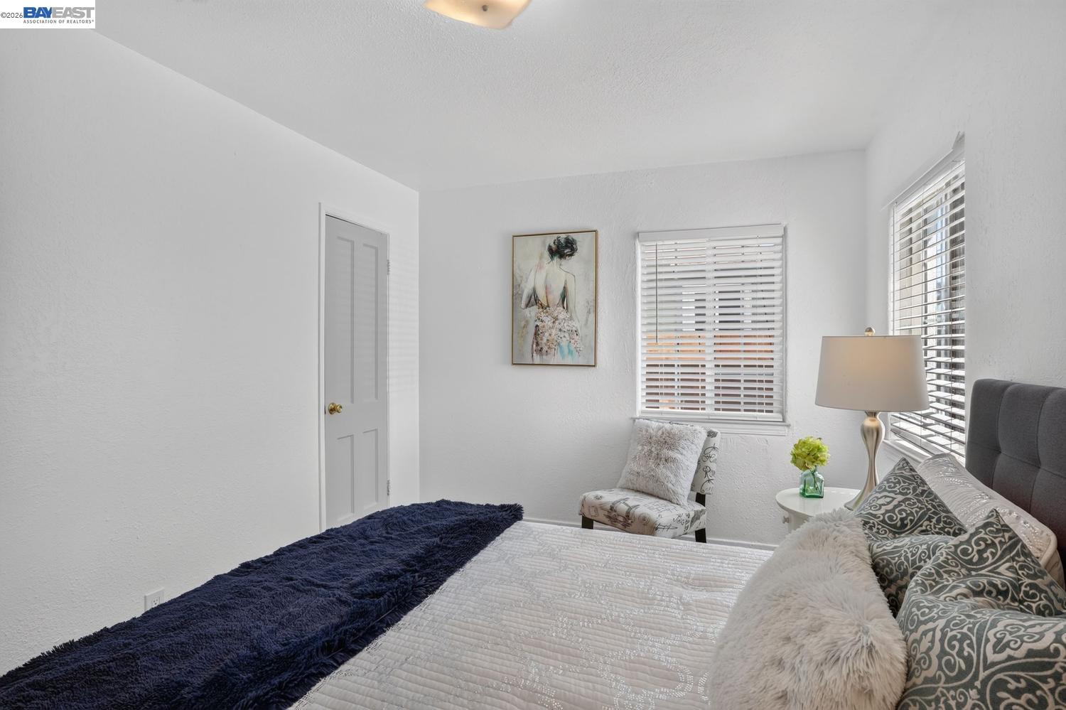 Bedroom with a white quilted bed, navy throw, and two windows with white blinds.