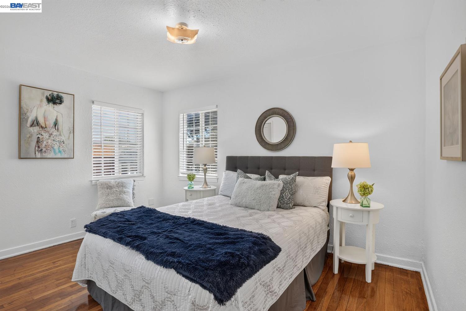 Staged bedroom with a white quilted bed, navy throw, hardwood floors, and two shuttered windows.