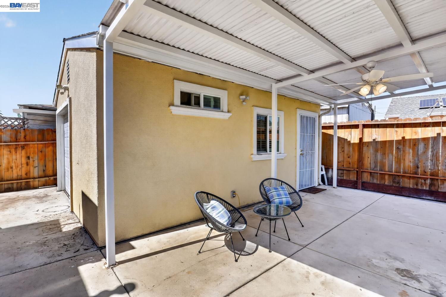 Covered backyard patio with two chairs, a ceiling fan, and wooden privacy fencing.