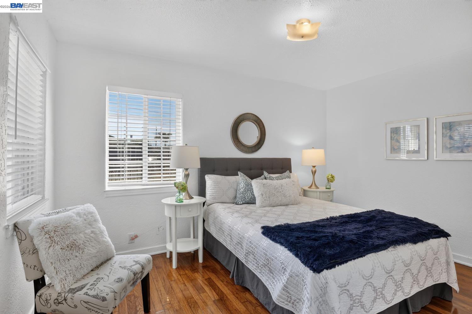 Furnished bedroom with a navy bed throw, hardwood floors, and a patterned chair by the window.