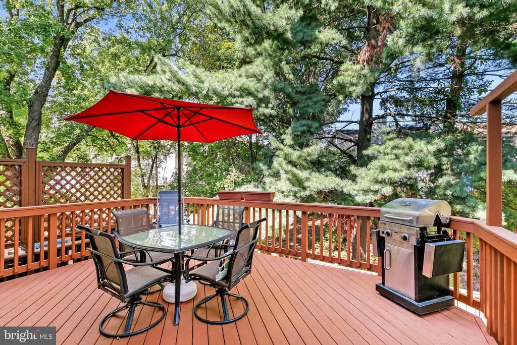 Outdoor deck with a red umbrella over a dining table and a stainless-steel grill, surrounded by trees.