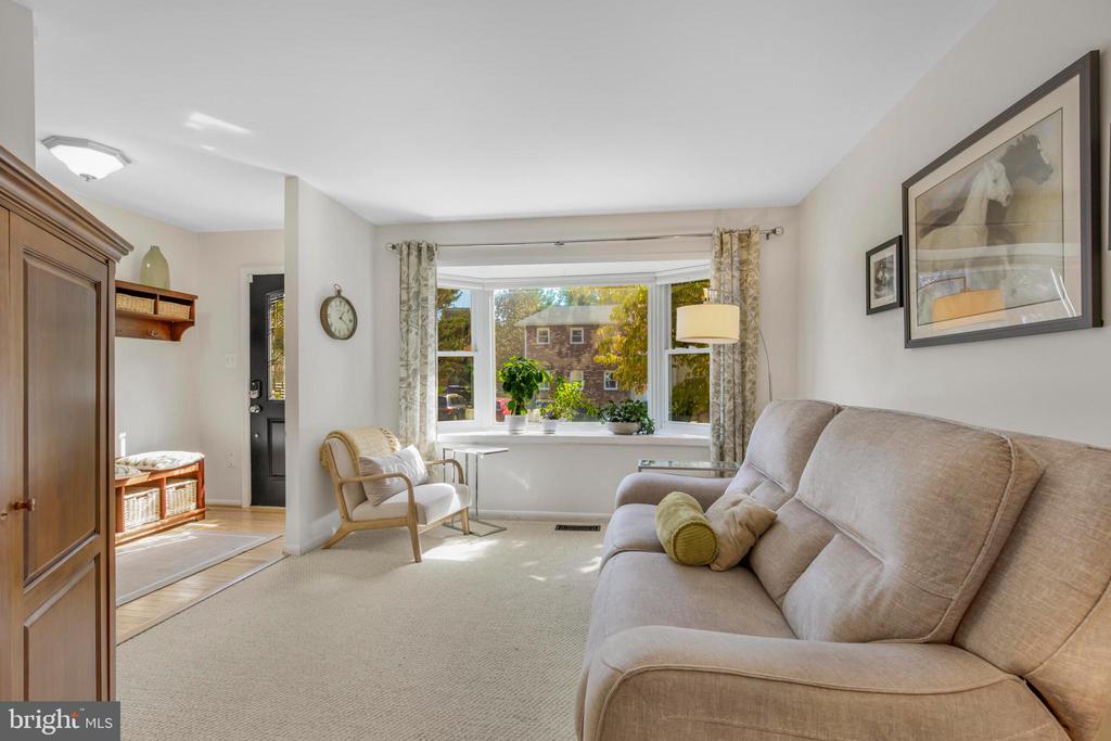 Living room with a beige sofa, armchair, and bay window overlooking trees and a brick building.