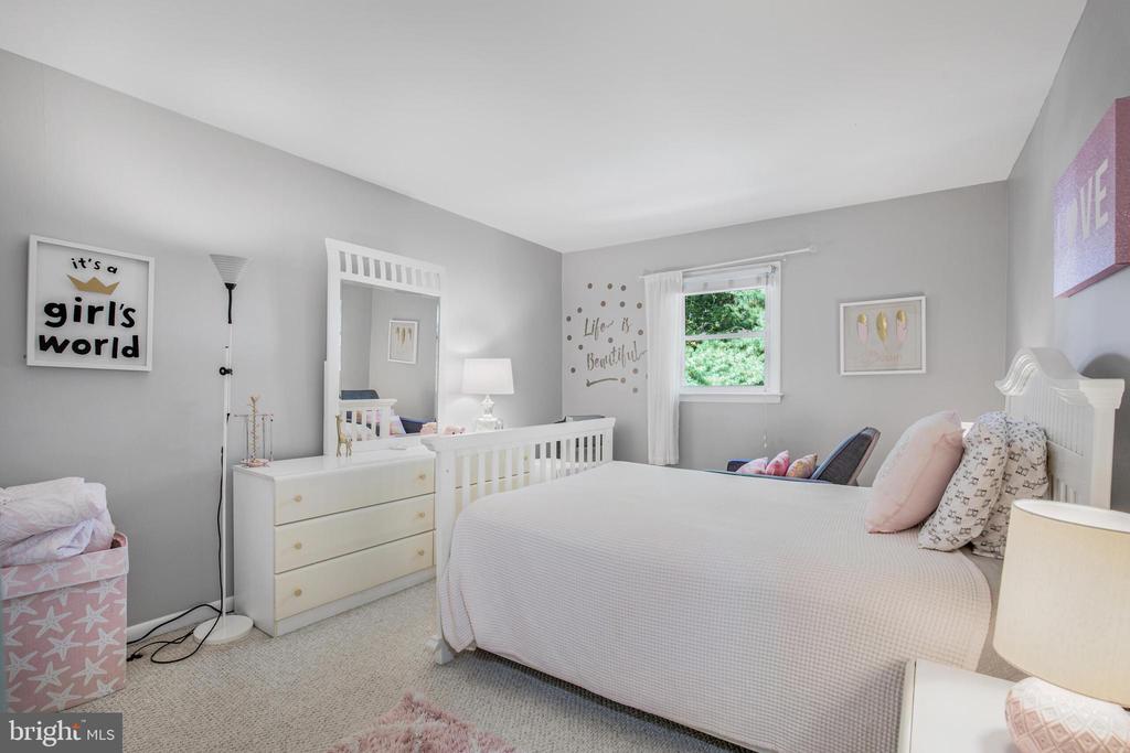 Bedroom with a large bed, white crib and dresser, and pink accents beside a window.