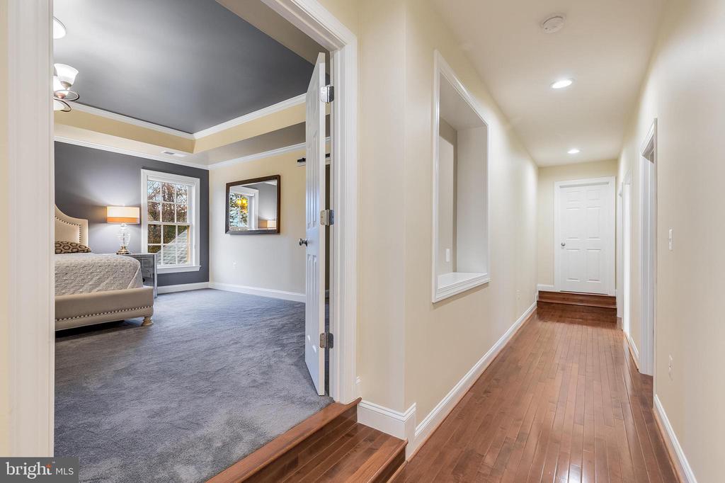 Hallway with hardwood floors beside a carpeted bedroom with a tray ceiling.