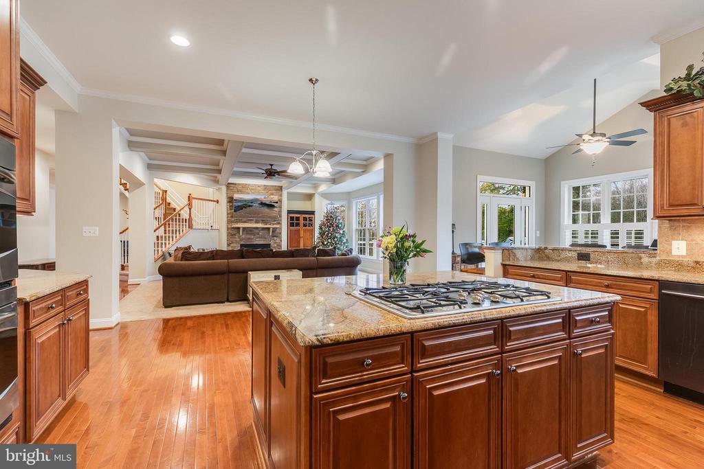 Open kitchen with a granite island and gas cooktop, looking toward a living room with a stone fireplace.