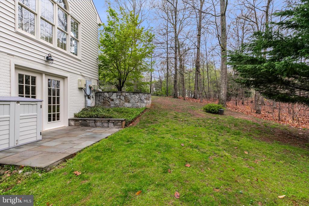 House exterior beside a paved patio, stone retaining wall, and wooded backyard.