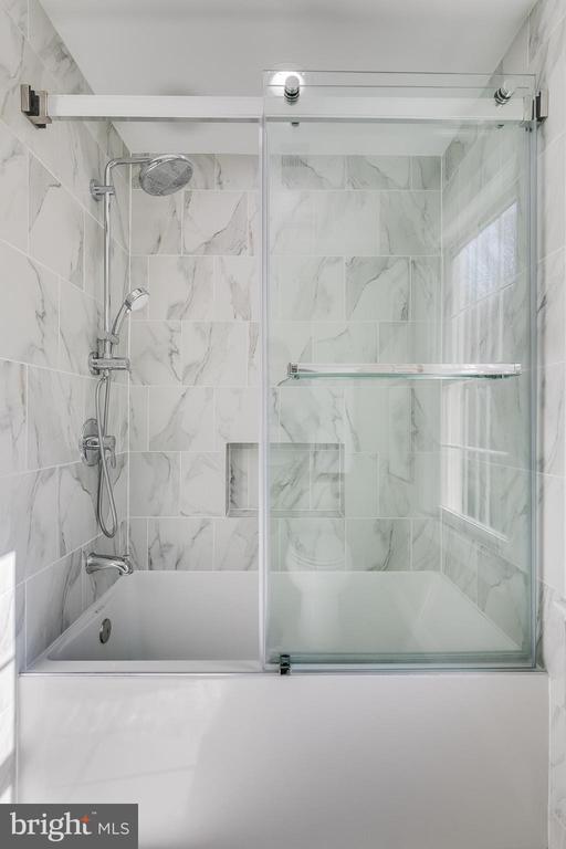 Bathroom with a glass-enclosed tub, marble-look tile, rainfall showerhead, and recessed niche.