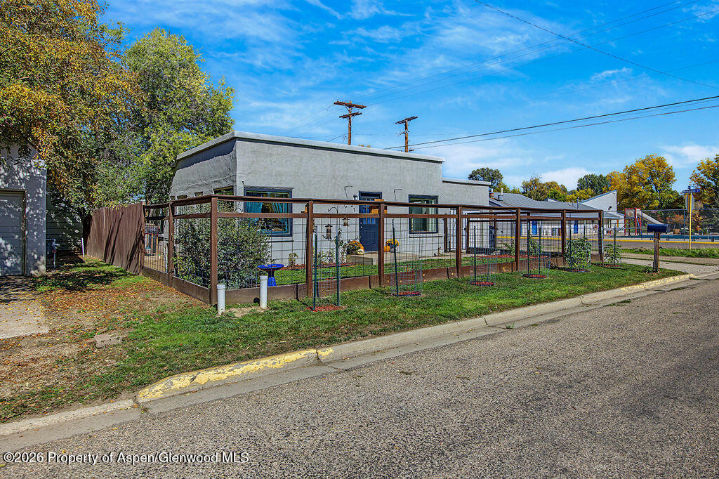 Gray single-story building behind a fenced garden, with autumn trees along the street.