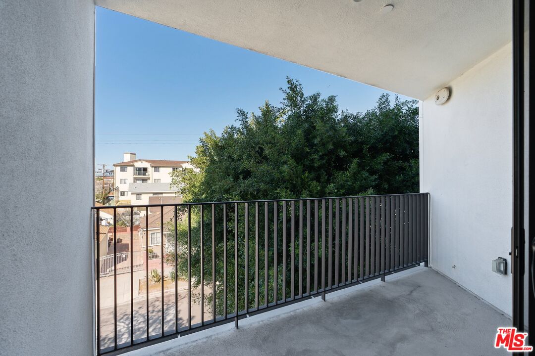 Covered balcony with black railing overlooking trees and neighboring buildings.